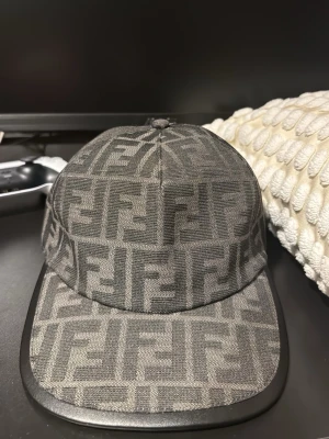 Svart Fendi keps med FF-mönster - Snygg svart keps från Fendi med det klassiska FF-logomönstret över hela kepsen. Kepsen har böjd skärm och är tillverkad i Italien. Materialet är en blandning av bomull och polyester, vilket ger en lyxig känsla och bra passform.