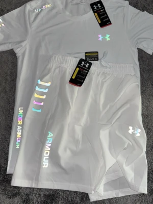  Under Armour - Snyggt vitt träningsset från Under Armour med t-shirt och shorts. Setet har reflekterande loggor och detaljer i regnbågsfärger. Tillverkat i lätt och snabbtorkande material som passar perfekt till träning eller löpning. Elastisk midja på shortsen för skön passform.