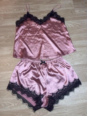 Rosa satinset med spetsdetaljer - Tvådelat set i glansig rosa satin med svarta spetsdetaljer. Linnet har tunna axelband och v-ringning med spetskant och shortsen har en liten rosett och matchande spets längs benen. I storlek S.