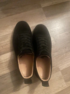 Svarta sneakers med röd sula - Snygga svarta sneakers från Christian Louboutin med klassisk röd sula och lågt skaft. Skorna har svarta skosnören och en stilren design i läder med beige innersula. Perfekta för dig som vill sticka ut med exklusiva detaljer. Kan gå ner vid en snabbaffär!! 