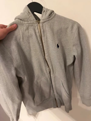 Grå hoodie från Polo Ralph Lauren - Klassisk grå hoodie med dragkedja från Polo Ralph Lauren. Tröjan har huva, två fickor framtill och den ikoniska mörkblå loggan broderad på bröstet. Tillverkad i mjuk bomullsblandning och har ribbade muddar vid ärmslut och nederkant.