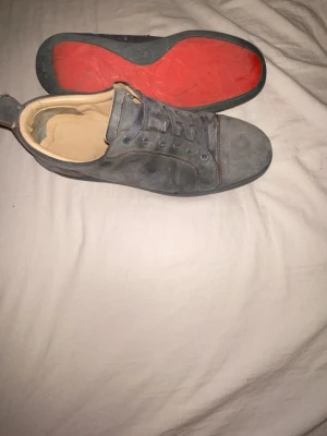Christian Louboutin - Gråa sneakers med lågt skaft och snörning. Skorna har en ovandel i mockaliknande material och en kontrasterande röd yttersula. Insidan är ljusbeige och designen är stilren och enkel, perfekt för en avslappnad look.