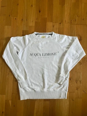 Vit sweatshirt från Acqua Limone - Säljer en vit sweatshirt från Acqua Limone med tryck på bröstet och en stor gul citron på ryggen. Tröjan har rund hals, ribbade muddar och är tillverkad i mjukt bomullsmaterial. Perfekt för en avslappnad och trendig look.