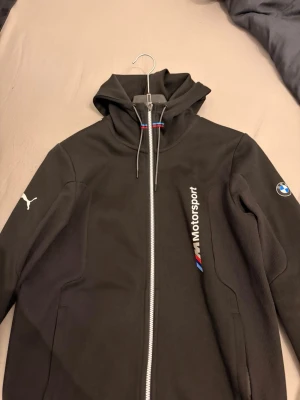 Svart BMW M Motorsport hoodie Puma - Säljer en svart hoodie från Puma i samarbete med BMW M Motorsport. Hoodien har dragkedja, huva med snören, vita och färgade detaljer samt loggor från både Puma och BMW. Tillverkad i mjukt material, perfekt för dig som gillar motorsport och streetwear.