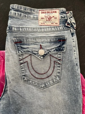 True Religion ljusblå jeans - Ljusblå jeans från True Religion med röda kontrastsömmar och ikonisk fickdesign. Modellen heter Becca. Snygga detaljer som logga på bakfickan. Säljer då de är alldeles för stora för mig!