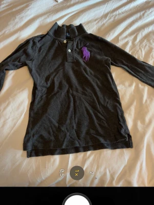 Svart långärmad piké från Ralph Lauren - Svart/grå långärmad pikétröja från Ralph Lauren med klassisk krage och två knappar framtill. Stor lila logga broderad på bröstet. Perfekt för en stilren och avslappnad look. Äkta 