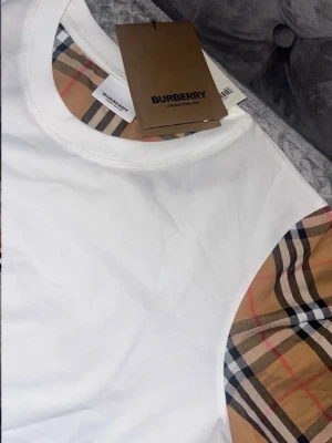Vit Burberry t-shirt med rutiga ärmar - Snygg vit t-shirt från Burberry med klassiska beige-rutiga ärmar. T-shirten har rund halsringning och den ikoniska Burberry-loggan på etiketten. Perfekt för dig som vill ha en stilren men ändå unik look.