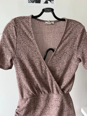 Rosa/lila omlottklänning med kort ärm - Kort rosa/lila omlottklänning med lätt melerat mönster och korta ärmar. Klänningen har v-ringning och rynkade detaljer i sidan för en snygg passform. Perfekt för dig som gillar en enkel men trendig stil.