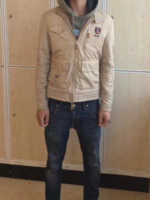 Beige vindjacka med patchdetaljer - Snygg beige vindjacka med dragkedja framtill, två stora fickor med knappar och coola patchdetaljer på bröstet. Jackan har en klassisk passform och är perfekt för dig som vill ha en stilren look. Passar till jeans och hoodie för en avslappnad vibe.