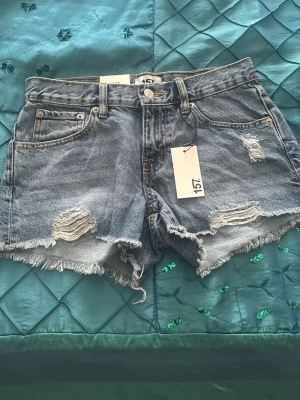 Blå denimshorts med slitningar 157 - Säljer ett par blå denimshorts från 157 med råa kanter och slitna detaljer framtill. Klassisk femficksmodell med låg midjad och knappgylf. Perfekta för sommaren och ger en avslappnad vibe till din outfit.
