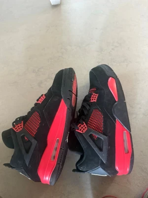 Svarta och röda Air Jordan 4 sneakers - Säljer ett par Air Jordan 4 sneakers i svart mocka med röda detaljer och meshpaneler. Skorna har klassisk siluett, svarta skosnören och synlig Air-enhet i sulan. Perfekta för dig som gillar streetwear och vill sticka ut med snygga färgkontraster.