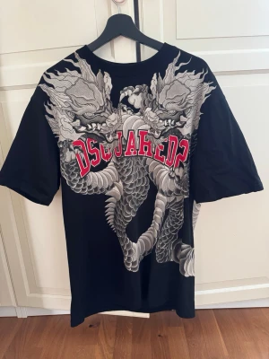 Svart Dsquared2 t-shirt med draktryck - Cool svart t-shirt från Dsquared2 med stort grått drakmotiv över ryggen och röd logga. T-shirten har korta ärmar och är gjord i mjuk bomull. Perfekt för dig som vill sticka ut med en unik och detaljerad design.