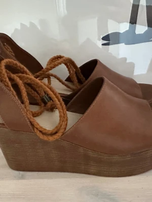 Bruna platåsandaler med snörning - Säljer ett par bruna platåsandaler med öppen tå och grov flätad snörning runt ankeln. Skorna har en hög platåsula i trälook och ovandel i brunt läder eller läderimitation. Perfekta för dig som vill ha extra höjd och en cool retrostil. Strl 39, använda en gång. 