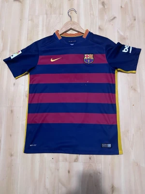 Barça matchtröja Nike 2015 med Neymar jr#11 tryck - Säljer en FC Barcelona matchtröja från Nike, säsongen 2015. Tröjan är blå med breda vinröda ränder, gula detaljer och klubbens emblem på bröstet. Materialet är lätt och andas, perfekt för fotboll eller träning. Autentisk märkning och sponsorloggor på ärmarna.