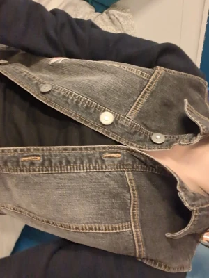 Svartgrå jeansjacka med kontrastsömmar - Cool svartgrå jeansjacka med klassiska kontrastsömmar och silverfärgade knappar framtill. Jackan har en tidlös design och passar perfekt till många olika outfits. 