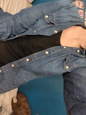Klassisk blå jeansjacka - Snygg blå jeansjacka med silverfärgade knappar och två bröstfickor med lock. Jackan har en rak passform och är tillverkad i klassiskt denimtyg. Perfekt att slänga över en t-shirt för en avslappnad look.