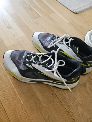 Svarta och vita Puma sneakers - Snygga sneakers från Puma i svart, vitt och grått med detaljer i gult. Skorna har mesh- och syntetmaterial, vita snören och en sportig design med låg profil. Perfekta för dig som gillar streetstyle och sportig look.