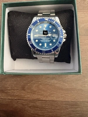 Rol*x Blå och silverfärgad Submariner-klocka - Snygg klocka i klassisk Submariner-stil med blå urtavla och silverfärgat armband i metall. Klockan har tydliga markeringar, datumfönster och vridbar bezel i blått. Perfekt accessoar för dig som vill sticka ut med en sportig och stilren look.