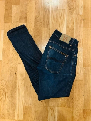 Nudie Jeans grim Tim - Snygga mörkblå jeans från Nudie Jeans med klassisk femficksdesign och kontrastsömmar. Jeansen har en smal passform och är tillverkade i denim med en tydlig vågdetalj på bakfickan. Perfekta för en stilren och avslappnad look.