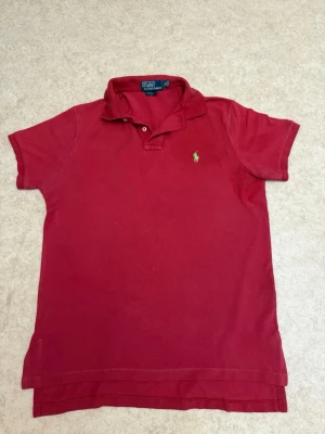 Röd pikétröja från Ralph Lauren - Klassisk röd pikétröja från Ralph Lauren med korta ärmar och krage. Tröjan har den ikoniska gröna broderade loggan på bröstet och är tillverkad i mjuk bomull. Perfekt för en avslappnad och stilren look.