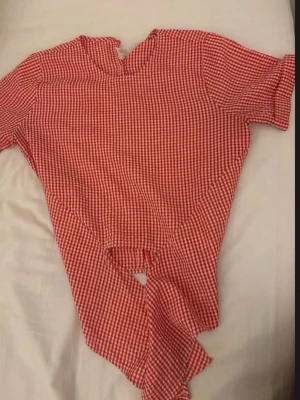 CROP TOP Röd- och vitrutiga blus med knyt - Trendig blus i röd- och vitrutigt mönster med korta ärmar och knyt framtill. Blusen har knappar längs ryggen och en liten ståkrage. Perfekt för en avslappnad men ändå stilsäker look. Materialet känns lätt och luftigt.