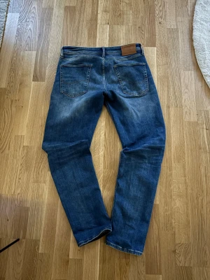 Blå raka jeans, Jack & Jones  - Säljer ett par klassiska blå jeans med raka ben och en liten slitning på benet som i mitt tyckte är ganska snyggt. Jeansen har fem fickor, normal midja och kontrastsömmar. Materialet är denim i bomull och de har en läderpatch bak i linningen.