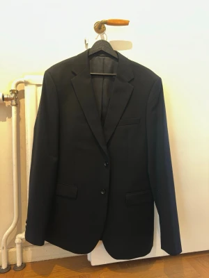 Svart kavaj från Dressmann Slim Fit - Stilren svart kavaj från Dressmann i modellen Slim Fit. Klassisk design med två knappar, spetsiga slag och tre fickor framtill. Perfekt för dig som vill ha en enkel och snygg kavaj till många olika tillfällen.