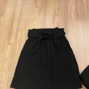 Svart kjol med knytband i midjan - Stilren svart kjol med hög midja och brett knytband som markerar midjan. Perfekt att matcha med en enkel topp eller stickad tröja.