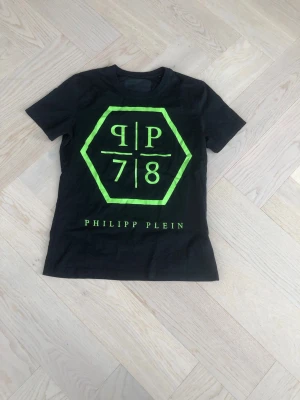 Svart Philipp Plein t-shirt neontryck - Snygg svart t-shirt från Philipp Plein med stort neon-grönt tryck framtill och logga. Klassisk rund hals och korta ärmar. Perfekt för dig som gillar streetwear och vill sticka ut med en unik design.