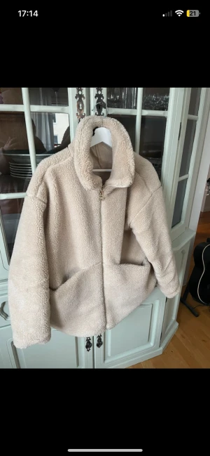 Beige teddyjacka med hög krage - Mysig beige teddyjacka med hög krage och dragkedja framtill. Jackan har två stora fickor och är tillverkad i fluffigt teddy-material som ger en avslappnad och trendig look. Perfekt för kyliga dagar när du vill vara både varm och snygg.