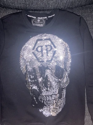Svart Philipp Plein tröja med paljettdödskalle - Snygg svart sweatshirt från Philipp Plein med en stor dödskalle i silverpaljetter på framsidan och PP-logga ovanför. Tröjan har rund hals och är tillverkad i mjukt material. Perfekt för dig som gillar statementplagg och streetwear.