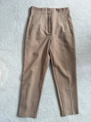 Beige kostymbyxa med hög midja - Stilrena beige kostymbyxor med hög midja och raka ben. Byxorna har pressveck framtill och två snedställda fickor. Tillverkade i ett mjukt, vävt material som ger en clean look. Perfekta för en dressad outfit.