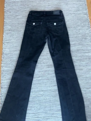Svarta bootcut jeans med fickdetaljer - Säljer ett par svarta bootcut jeans med snygga fickdetaljer bak och silverfärgade knappar. Jeansen har låg midja och klassisk femficksmodell. Perfekta för dig som gillar en retroinspirerad look och vill ha ett par jeans som sticker ut.