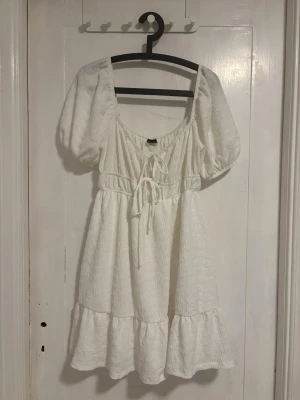 Vit kort klänning med puffärm - Superfin vit kort klänning med puffärmar och knytning framtill. Klänningen har en volang nertill och är tillverkad i ett strukturerat bomullstyg. Perfekt för sommarens alla tillfällen och ger en romantisk vibe.