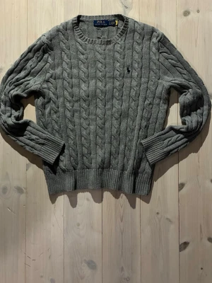 Grå kabelstickad tröja Polo Ralph Lauren - Kabelstickad grå tröja från Polo Ralph Lauren med rund halsringning och ribbade muddar. Klassisk design med diskret broderad logga på bröstet. Tillverkad i mjukt material som passar perfekt för kyligare dagar.