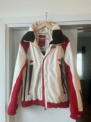 Vit och röd Åre 2007 Skid jacka från Halti - Sportig vindjacka från Halti i vitt med röda och svarta detaljer. Jackan har dragkedjor framtill, hög krage och justerbara muddar. Märkt med Åre 2007 och officiell leverantör till FIS Alpine World Ski Championships. Perfekt för dig som gillar en aktiv stil.
