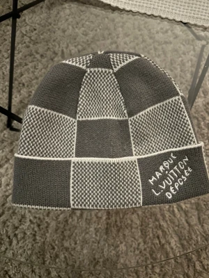 Gråvit rutmönstrad mössa  - Snygg mössa i grått och vitt med rutmönster. Stickad design med broderad text 'Marque L. Vuitton Déposée' på kanten. Perfekt accessoar för dig som gillar streetstyle och exklusiva märken.