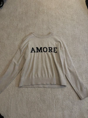 Beige tröja med svart text AMORE - Snygg beige tröja med svart texten 'AMORE' framtill. Tröjan har rund halsringning och långa ärmar. Tillverkad i mjukt material som känns skönt mot huden. Perfekt för dig som gillar enkel men trendig stil.