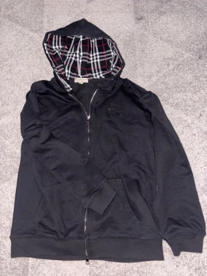 Svart hoodie från Burberry - Svart hoodie från Burberry med dragkedja och klassiskt rutigt foder i huvan. Tillhörande svarta snören med metalländar där Burberry-loggan syns. Hoodien har ribbade muddar och är tillverkad i mjukt bomullsmaterial. Perfekt för en stilren och avslappnad look.
