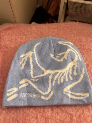 Ljusblå mössa från Arc'teryx - Snygg ljusblå mössa från Arc'teryx med vitt grafiskt mönster och broderad logga framtill. Perfekt accessoar för kalla dagar, tillverkad i mjukt stickat material som sitter skönt på huvudet.
