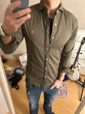 Olivgrön skjorta från Ralph Lauren - Snygg olivgrön skjorta från Ralph Lauren med klassisk krage och knappar framtill. Skjortan har lång ärm och den ikoniska broderade loggan i gult på bröstet. Perfekt till jeans för en avslappnad men stilren look.