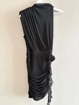 Svart asymmetrisk kort klänning - Snygg svart kortklänning med draperad design och asymmetrisk axel. Klänningen har en dekorativ blomdetalj i sidan och rynkade partier längs kjolen. Tillverkad i ett mjukt, stretchigt material som ger en smickrande passform.