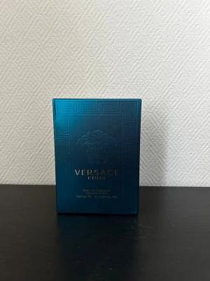 Versace Eros Eau de Toilette 100 ml - Versace Eros Eau de Toilette i en lyxig, mörkblå kvadratisk glasflaska med gulddetaljer och Medusa-huvudet på framsidan. Flaskan rymmer 100 ml och har en elegant, metallisk blå och guld kork. Perfekt för dig som gillar exklusiva och ikoniska dofter.