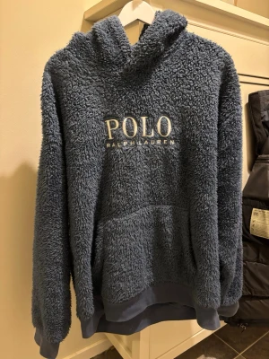 Blå teddyhoodie från Polo Ralph Lauren - Mysig blå teddyhoodie från Polo Ralph Lauren med stor broderad logga på bröstet. Hoodien har huva, magficka och ribbade muddar vid ärmslut och nederkant. Tillverkad i fluffigt, mjukt material. Riktigt skön och inte mycket använd. Storlek/ L/G