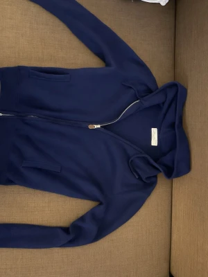 Mörkblå hoodie från Sayless i cashmere - Mörkblå hoodie från Sayless i mjuk cashmere. Tröjan har dragkedja framtill, två fickor och en klassisk huva. Perfekt för dig som vill ha en stilren och bekväm hoodie med exklusiv känsla.