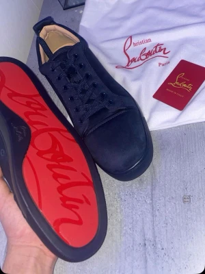 Mörkblåa Christian Louboutin sneakers - Snygga mörkblåa sneakers från Christian Louboutin med klassisk röd sula och diskret logga undertill. Skorna har en låg profil, rund tå och är tillverkade i mocka med matchande mörkblåa skosnören. Perfekta för dig som gillar exklusiv och stilren design.