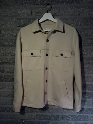 Beige overshirt från Zara - Stilren beige overshirt från Zara med klassisk krage och svarta knappar. Två stora bröstfickor med lock och knapp samt två sidofickor. Overshirten har en rak passform och är tillverkad i ett mjukt material som ger en clean look. Storlek S men sitter som en medium. OBS det är velourtyg. Aldrig använd.