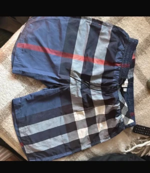 Rutiga badbyxor i blått och rött - Snygga badbyxor med klassiskt rutmönster i blått, svart, vitt och en röd linje. De har elastisk midja och är tillverkade i ett lätt, snabbtorkande material. Perfekta för stranden eller poolen.