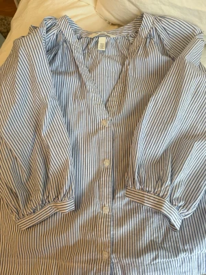 Blårandig blus från H&M med puffärm - Snygg blårandig blus från H&M med knäppning framtill och puffiga trekvartsärmar. Tillverkad i ett lätt bomullstyg som känns luftigt och fräscht. Perfekt till jeans eller kjol för en trendig look.