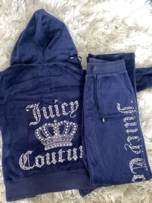 Blå velourset från Juicy Couture - Mörkblått velourset från Juicy Couture med hoodie och byxor. Setet har glittrande strasstenar med loggan 'Juicy Couture' och en krona på ryggen samt text på benet. Mjuk och bekväm kvalitet, perfekt för chill dagar hemma. Byxorna är lågmidjade❤️
